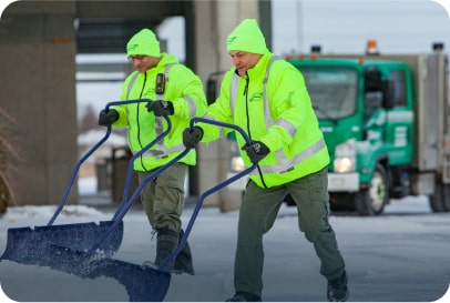 Two Clintar employees in bright neon jackets and hats shovel snow on a road in an industrial area.