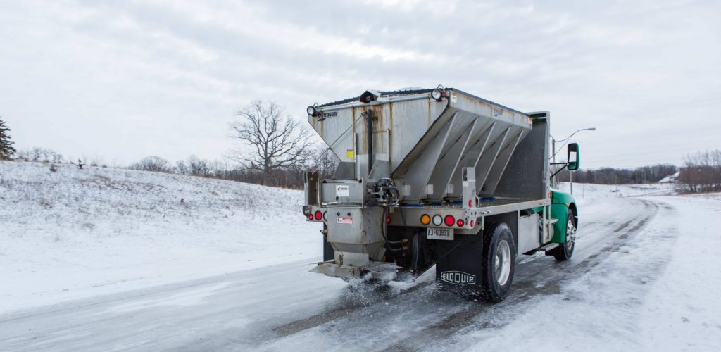 A Clintar truck with a large metal spreader is efficiently salting the snowy and icy road, assisting in snow clearing.