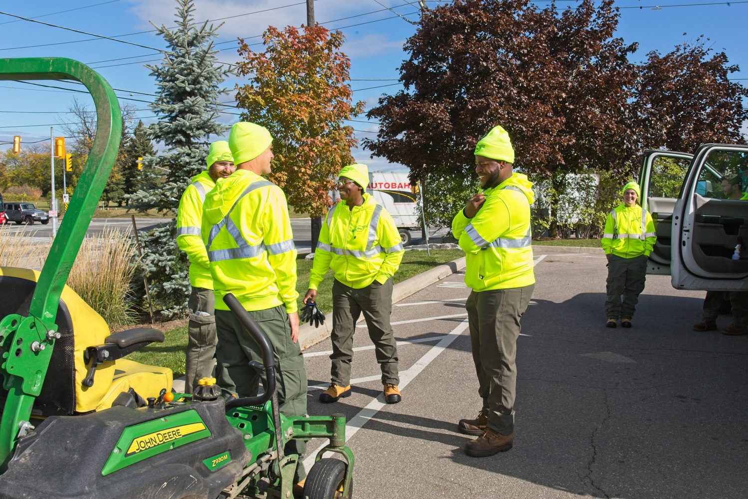 Six smiling Clintar employees in bright yellow safety jackets stand in a parking lot near a parked vehicle and landscaping equipment.